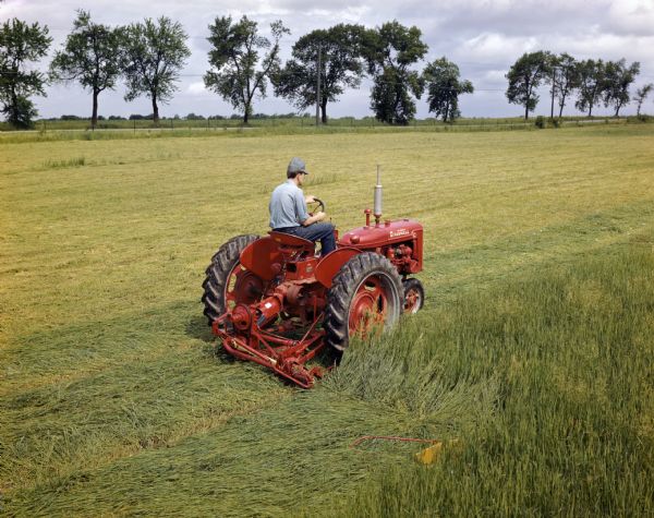 Elevated view of a farmer in a field operating a McCormick Farmall C tractor with attached mower.