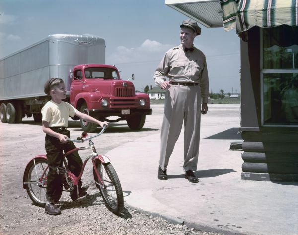 A uniformed driver of an International semi-truck (tractor-trailer) is talking with a boy on a bicycle outside a restaurant or diner.