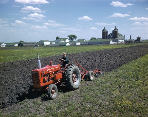Three-quarter view from front left towards a farmer in a field operating a McCormick standard diesel WD-6 tractor with plow. Farm buildings are in the background.