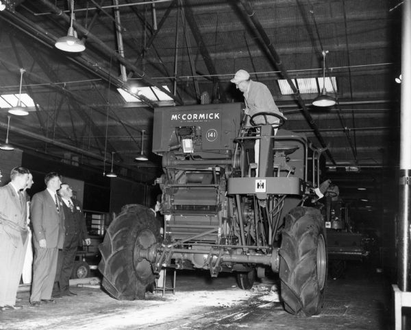 Tour of International Harvester's East Moline Works (factory) for the St. Cloud combine promotion. Original caption reads: "There are hundreds of inspections on the combine assembly line - and before that there are many other in foundry, forging and stamping operations, the visitors learned. Here they see the clutch on the No. 141 combine engine being tested and inspected. This is one of the last tests on the assembly line. Feeder has been removed from this machine for additional testing and inspection."
