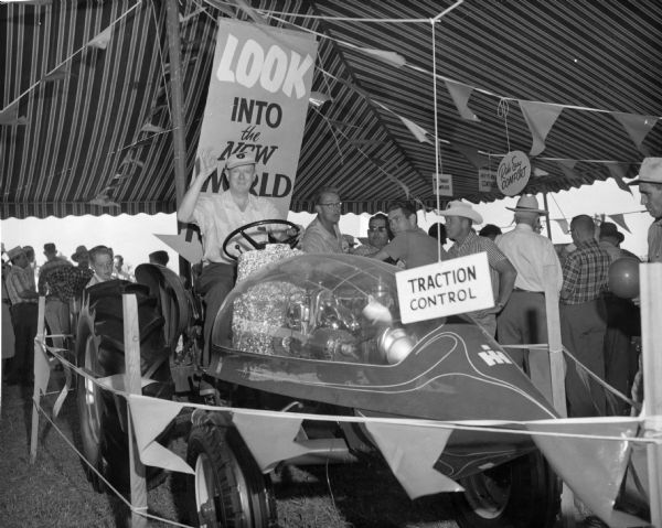 Men and a boy examining a prototype "Tractor of the Future" in an exhibit during the St. Cloud combine promotion. Signs hanging around the display read: "Look into the New World" and "Traction Control." A man is saluting from the seat of the futuristic tractor.
