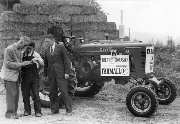 First Farmall M tractor produced at International Harvester's Doncaster Works (factory) in England, Great Britain. Original caption reads: "First Doncaster M Tractor being delivered to Alan Neale, son of Arthur Neale former Chairman of the board." A large stack of hay bales is in the background.
