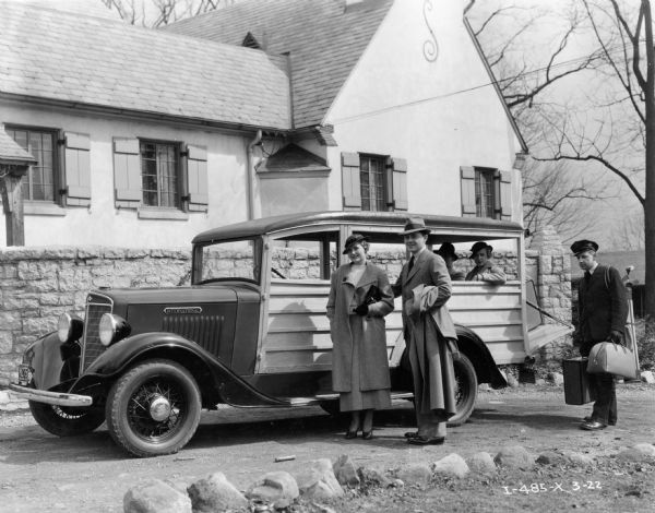 A man and a woman prepare to board an International station wagon ("woody") while the uniformed driver handles their luggage. Another man and woman are already sitting in the vehicle. The station wagon is parked outside a house or small hotel.