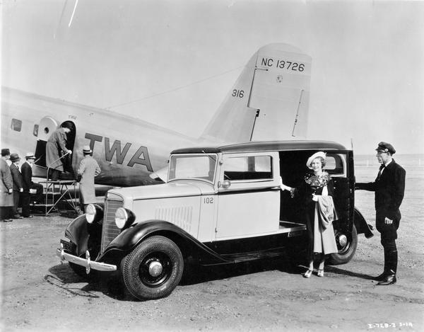 A woman is stepping out of an International station wagon while the driver is holding the vehicle's door open. The station wagon is parked near the tail section of a TWA airplane. Passengers are boarding the plane.