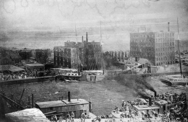 Elevated view of men and ships near the McCormick Works and the Chicago River during the Chicago Fire of 1871. The original caption reads:  "Reproduction of photograph taken during the big Chicago fire, 1871.  Photograph taken from south bank of river, east of Rush Street, showing McCormick Works in the center on the north bank of the river." The McCormick factory was ultimately destroyed in the fire.