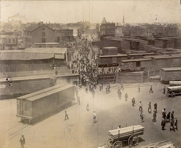 Elevated view of factory workers entering the Oakley Avenue gate of the McCormick Reaper Works. The men are filing in from the street, passing railroad boxcars, wagons and stacks of lumber.