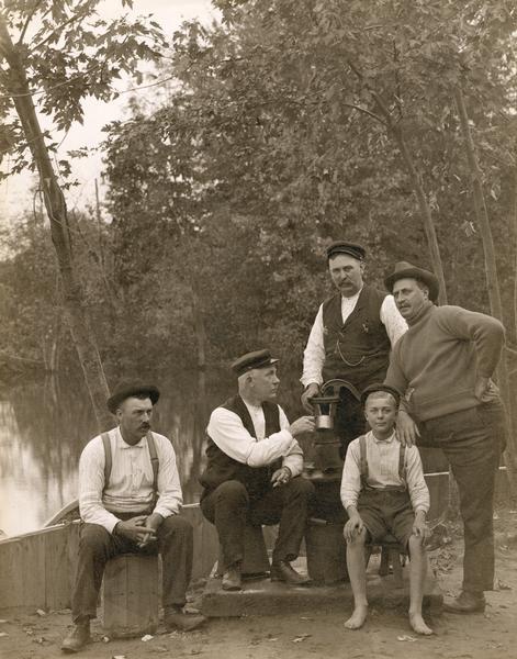 Group of men and a boy gathered near a body of water. The men are sitting around a water pump.