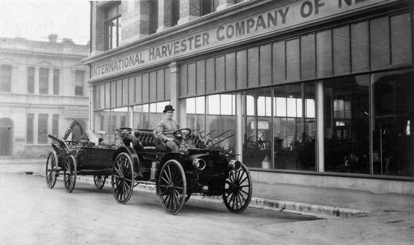 M.M. Black is sitting in an International Auto Buggy attached to a trailer full of agricultural implements (or parts) in front of the Christchurch office of the International Harvester Company of New Zealand. The original caption reads: "Mr. M. M. Black in front of Office at Christchurch, New Zealand."