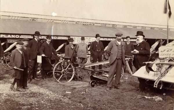 Two boys and a group of men inspect Plano mowers at a fairgrounds in Cork, Ireland. At the time, Plano was one of the brand names sold by International Harvester. The original caption reads:  "Mr. McBride, Agent at Cork for Plano is the old gentleman with the beard near the Jones Lever Binder, writing an order.  Fred W. Jones is the first man at the left of line next to the two boys."