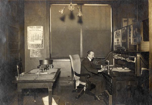 J.W. Wischart working in the office of the International Harvester general agency building in Fort Wayne, Indiana. The office contains a large desk, calendars, telephone, and other office equipment.