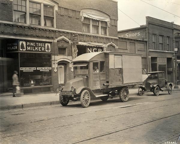 International Model 31 truck parked on an urban street in front of a Pine Tree Milker store and a National Cash Register store. A woman is walking by on the sidewalk.