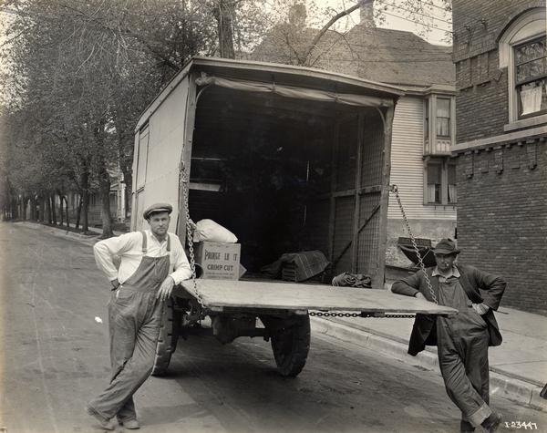 Two men pose next to the open back of an International delivery truck.  A box of pipe and cigarette tobacco sits in the back of the truck. The truck may be a Model 31.