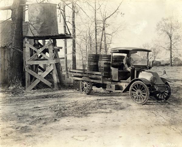 Man hauling barrels of water(?) in the back of an International motor truck. The truck may be a Model F or Model H. The truck appears to be pulling away from a water tower.