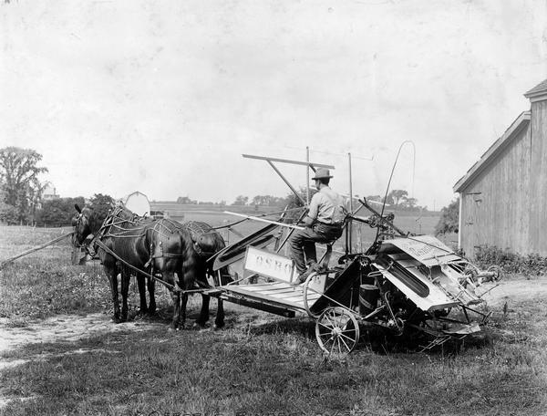 Farmer driving a horse-drawn Osborne grain binder near a barn.