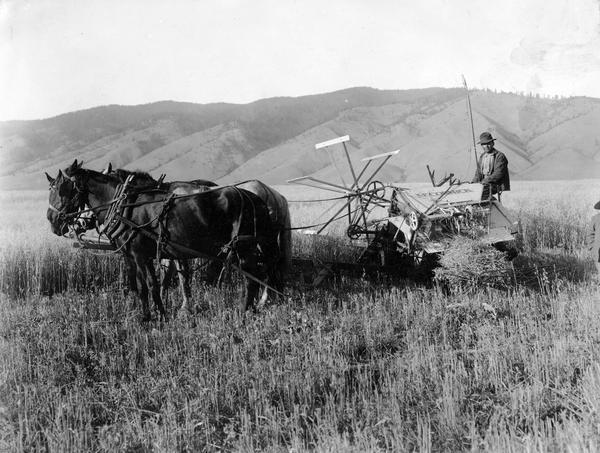 Man operating a horse-drawn McCormick grain binder against a mountainous backdrop.