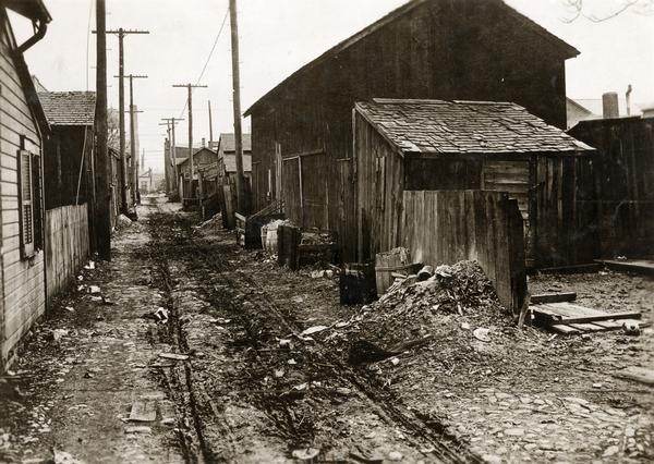 Squalid, littered alley. Tire tracks run through the mud between wooden buildings.