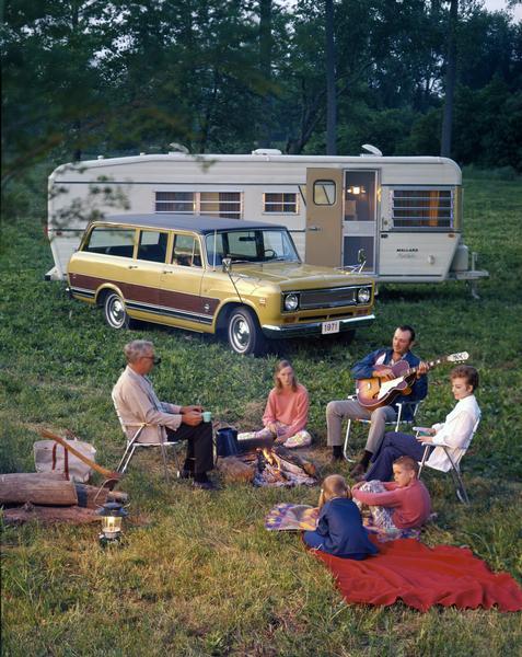 Color advertising photograph of a family enjoying a campfire near their Travelall 1010 truck and Mallard camper. The father of the family strums his guitar, while the others listen.