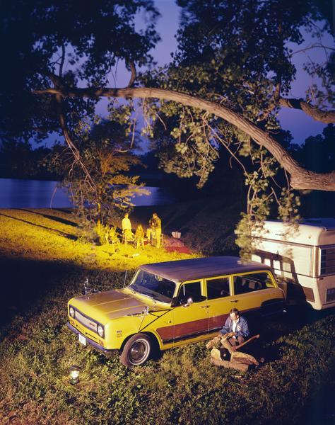 Color advertising photograph of a family singing by a campfire near a Travelall 1010 truck and a Mallard camper. A man is in the foreground chopping wood for the fire.