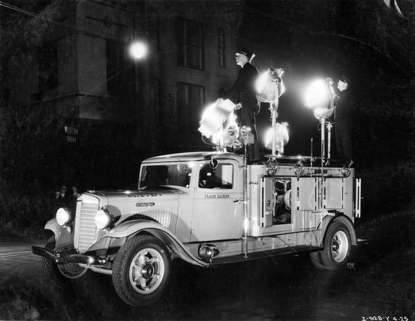 International Model C-40 fire truck with floodlights. The truck was owned by the Memphis, Tennessee fire department. A man stands on top of the truck and guides a flood light to a nearby building.