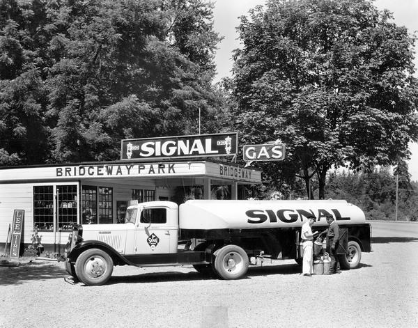 International oil truck delivers to a Bridgeway Park gas station. Original caption reads: "International equipped with a tank body of 1,950 gallon capacity owned by E.C. Mennis of Salem, Ore., who has the distribution of Signal Oil Co. products in the northern portion of the Willamette Valley in Oregon. This truck serves a number of the minor dealers in that territory and covers as much as 100 miles per day."