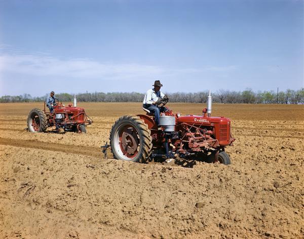 Men are operating McCormick Farmall tractors with seeding attachments. One tractor is a Farmall C and the other is a Farmall H.