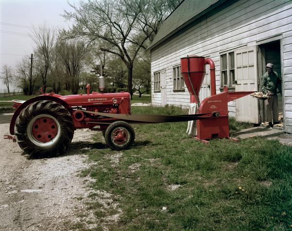 View of a man operating a hammer mill powered by a McCormick Super W-4 tractor in the doorway of a barn.