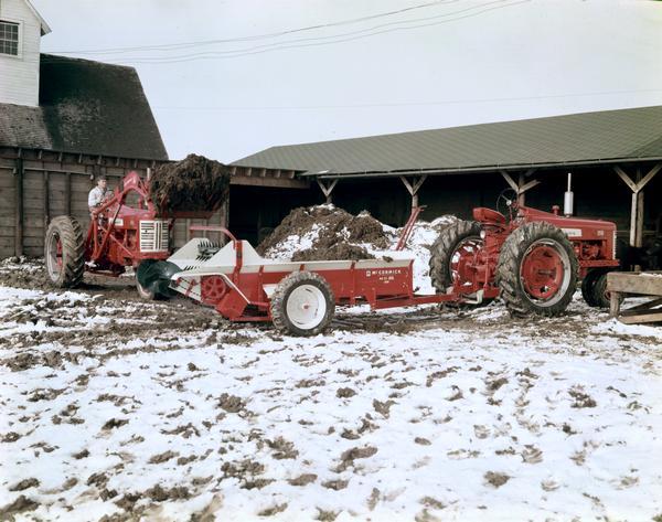 A man is using a Farmall 450 tractor to load a McCormick No. 21 manure spreader that's attached to a Farmall 350 tractor in a snow-covered farm yard.
