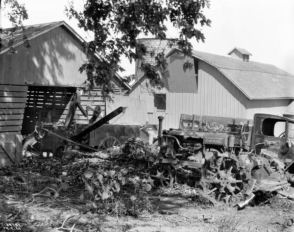 Two farmers shelling corn with a belt-driven McCormick corn sheller powered by a Farmall Regular tractor. Also in the farm yard are a McCormick-Deering wagon and an International truck.