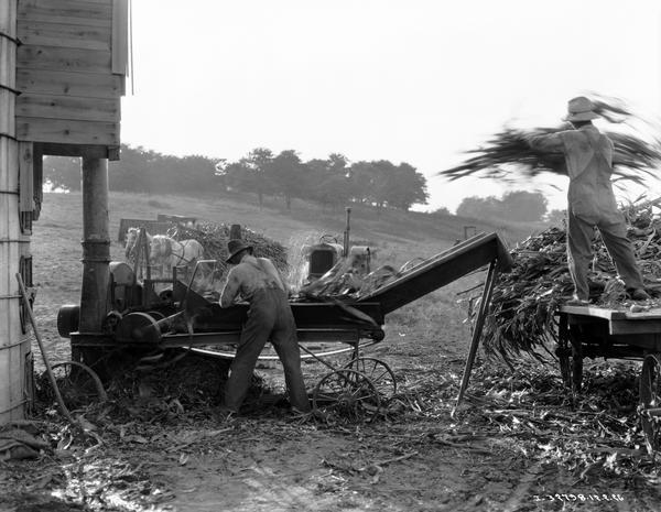Two farmers feeding corn stalks into an International Harvester ensilage cutter. In the background is a McCormick-Deering tractor.