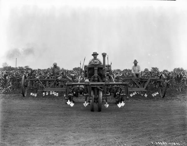 View from front of three farmers operating a McCormick-Deering tractor with shovel-type cultivators near a cornfield.