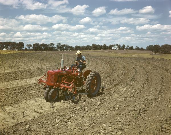 View towards a man in a field operating a McCormick Farmall C tractor with front-mounted cultivator. The color photograph was taken either at or near the company's experimental farm.