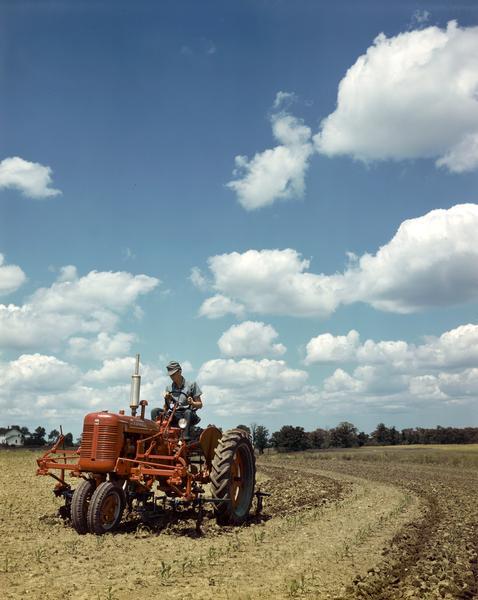 View towards a man in a field operating a McCormick Farmall C tractor with front-mounted cultivator against a bright blue sky strewn with fluffy white clouds. The color photograph was taken either at or near the company's experimental farm in Hinsdale, Illinois.