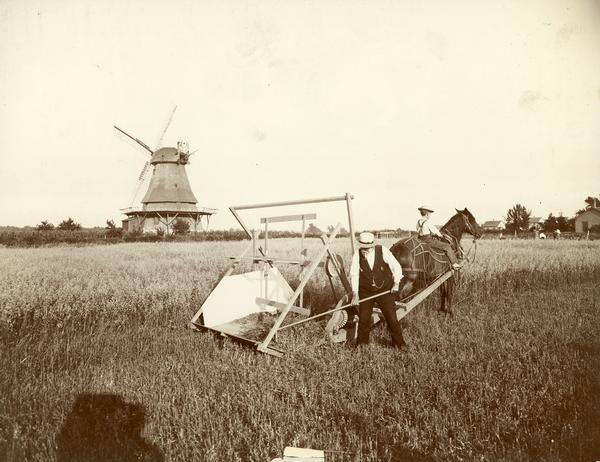 Man and a boy working with a horse-drawn reaper in a field. The reaper is likely a model or replica of the McCormick reaper of 1831. A windmill is in the background and the photographer's shadow is in the foreground. The photograph may have been taken for the McCormick Harvesting Machine Company or its successor, the International Harvester Company.