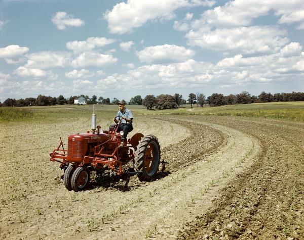 View towards a farmer in a field riding a McCormick Farmall C tractor with front-mounted cultivator. Taken either at or near the company's experimental farm.