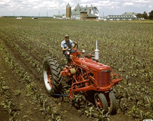 Slightly elevated view of a man in a field operating a McCormick Farmall H tractor with a mounted cultivator. A barn with "Hinsdale Farms" painted on the front is in the background.