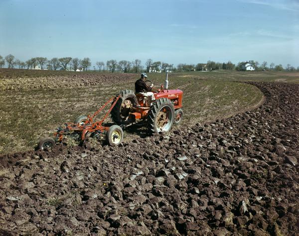 View towards a man in a field operating a McCormick Farmall H tractor with attached plow, either at or near the company's experimental farm in Hinsdale.