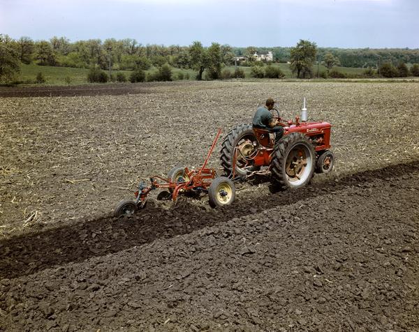Slightly elevated view of a man in a field operating a Farmall H tractor and plow, either at or near the company's experimental farm in Hinsdale, Illinois.