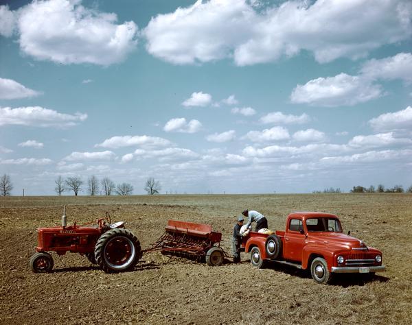 Farmers are unloading bags of seed from the back of an International L-series pickup truck in a field. A McCormick Farmall H tractor with attached grain drill is parked nearby, either at or near the company's experimental farm in Hinsdale.