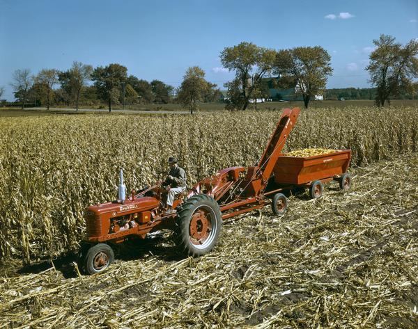 Slightly elevated view of a man picking corn with a McCormick Farmall H tractor, a McCormick 14-P pull-type corn picker and a wagon. The photograph was taken at or near the company's experimental farm in Hinsdale, Illinois.