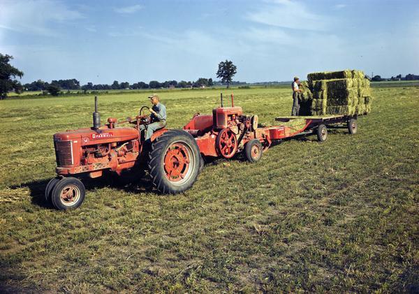 Slightly elevated view of a man driving a McCormick Farmall M tractor, and a man baling hay with a power hay baler, and trailer. The baler is powered by an International power unit.