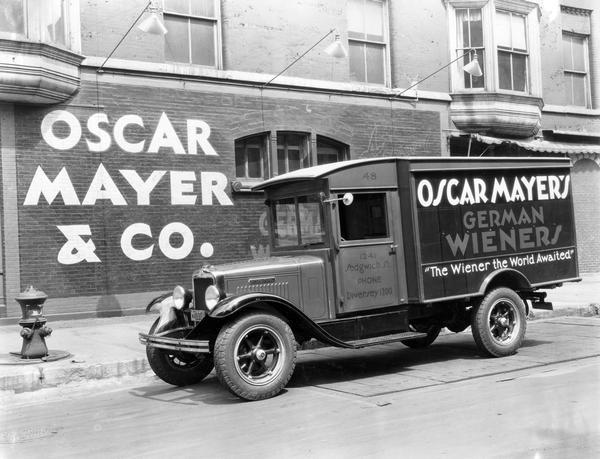 Oscar Mayer delivery truck parked outside an Oscar Mayer & Co. building. The truck is an International AW-2 and features 32 x 6 tires. The side of the truck includes the words: "Oscar Mayer's German Wieners - 'The Wiener the World Awaited.'"