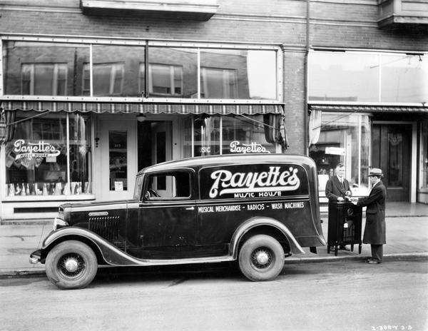 Two men are standing at the back of an International Model C1 panel truck owned by Payette's Music House in Aberdeen, Washington. The men are standing with a new radio in front of Payette's store.