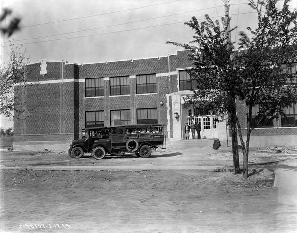 Two buses with children on board leaving a school in Quinter, Kansas.  One is an International bus serving Quinter Consolidated schools. Three people are standing near the front door of the school.