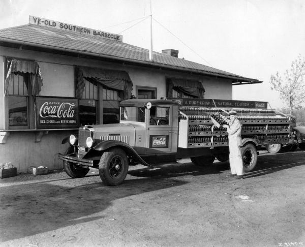 Delivery man unloading Coca-Cola (soda) bottles from his International A-4 truck with steel bottlers body. The truck was owned by the Midwest Dairy Company and is parked outside a restaurant called "Ye-Old Southern Barbecue."