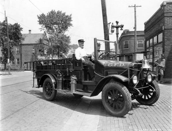 Two fire fighters are sitting in an International fire truck on a street. Another man is standing on the back of the truck. On the side of the truck is a painted sign that reads: "C.F.D." In the background on the right is a man leaning on the corner of a commercial building.