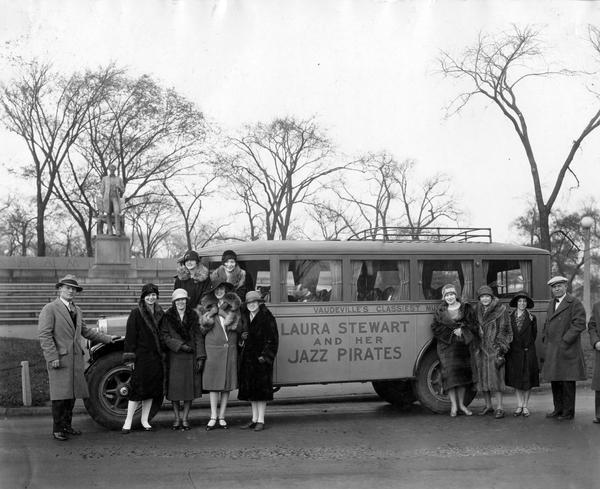 Laura Stewart and her Jazz Pirates pose alongside an International bus. The bus features windows with curtains and luggage rack(?) on top. There is a monument in the background.