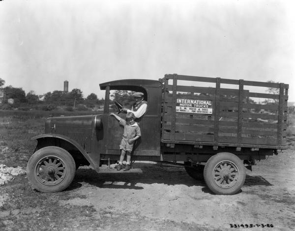 Portrait of a man sitting in the driver's seat of a truck, and a girl standing on the running board. The sign on the truck reads: "H.M. Reeve and Sons, East Moriches."