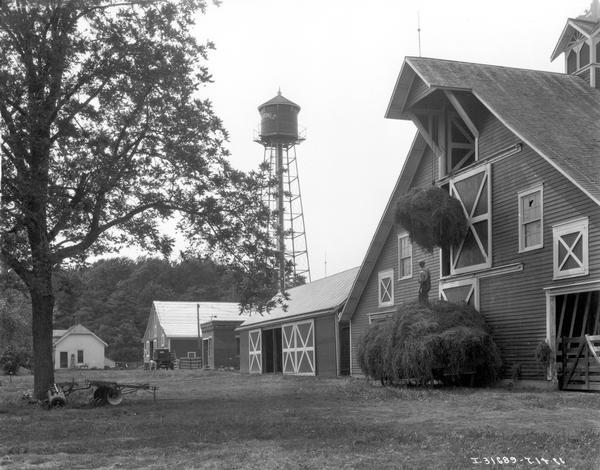 Farmer loading hay from a wagon into a large barn using a winch. There are other farm buildings, a water tower, and a farmhouse in the background.
