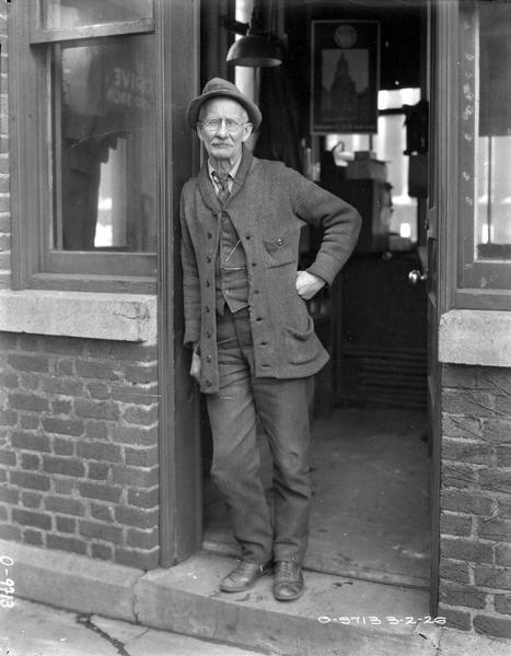 Factory employee standing in an office doorway (possibly a gate house?) at International Harvester's Osborne(?) Works. He is wearing eyeglasses, and a sweater, necktie, vest, hat, and trousers.