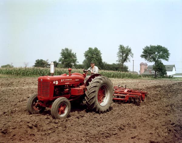 Three-quarter view towards front left of a farmer plowing a field with a McCormick Super WD-9 tractor and an attached disk plow.